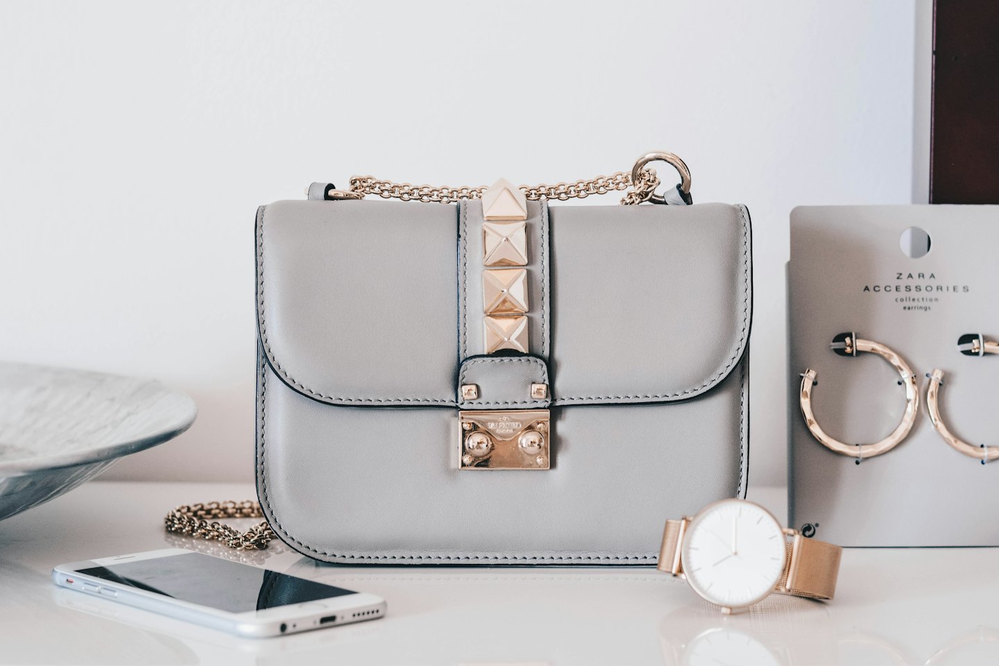 Graue Handtasche, Smartphone, Uhr und Creolen auf weißem Untergrund.