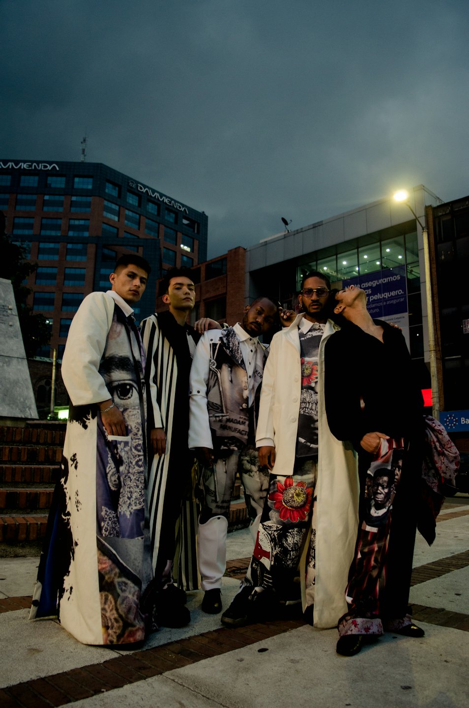 Fünf junge Männer in stylischen Trenchcoats mit grafischem Aufdruck posieren in der Abenddämmerung auf einer Stadtstraße.